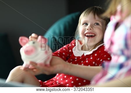 Portrait Of Beautiful Blonde Girl Resting On Sofa Indoors. Cute Kid Saving Up Money In Piggy Bank. M