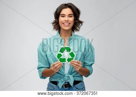eco living, environment and sustainability concept - portrait of happy smiling young woman in turquoise shirt holding green recycling sign over grey background