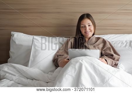 Beautiful Girl In Bathrobe Studies Online With Tablet Sitting In Soft Bed In Hotel Room During Self-