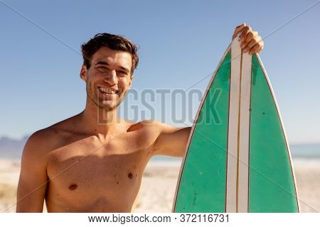 Caucasian man enjoying his time at the beach with his friends on a sunny day, standing shirtless, holding a surfboard and looking at the camera