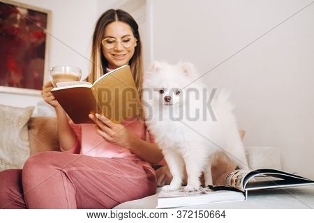 A Girl In Pajamas At Home Reads A Book With Her Dog Spitzer, The Dog And Its Owner Are Resting On Th