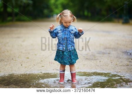 Child Wearing Red Rain Boots And Jumping In Puddle On A Summer Day. Adorable Toddler Girl Having Fun