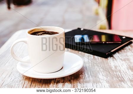 Coffee Cup And Smart Phone Outdoors On A Table At Coffee Shop Or Cafe With Blurred Background