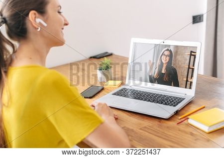 A Young Woman Sits Indoor And Using App On Laptop For Video Call To Female Coworker, Colleague Or Fr