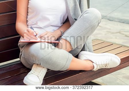 Smiling Young Woman Wearing Glasses Making Notes In Notebook While Sitting On The Beanch Outdoors. L