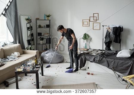 Young man sweeping floor at home after party: bottles, crumps and poker chips on floor