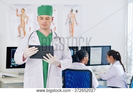 Serious Surgeon Reading Medical History Of Patient On Tablet Computer, His Colleagues Discussing Wor