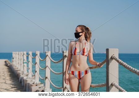 Young Woman In Bikini Standing At The Beach And Wearing Black Face Mask. Public Beach Rules During C