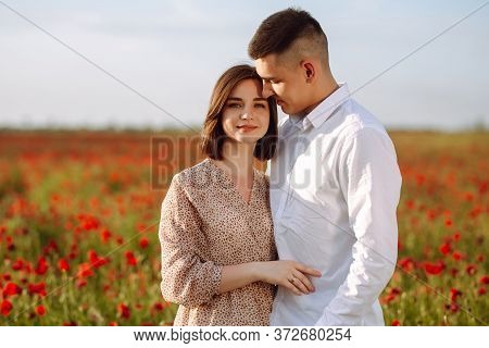 Young Romantic Couple Standing On The Red Poppy Field On A Sunny Day Showing Love To Each Other. Hap