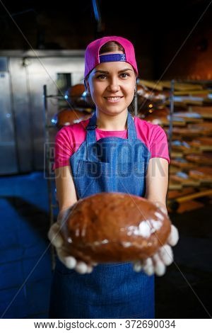 A Baker Girl Takes Hot Bread In A Bakery Against The Background Of Shelving With Bread. Industrial P