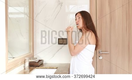 Long Haired Brunette In White Towel Brushes Teeth With Toothbrush Looking Into Bathroom Mirror At Ho