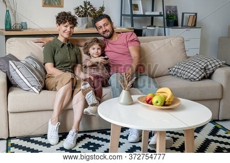Cheerful family of three in casualwear looking at you while sitting on large comfortable couch with cushions in living-room by small table