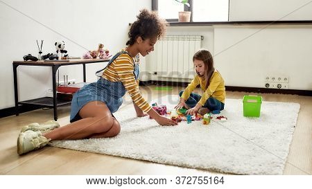 African American Woman Baby Sitter Entertaining Caucasian Cute Little Girl. They Are Playing With To