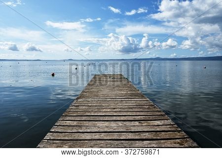 Wooden Jetty For Boats Against A Blue Sky With Some Clouds. Floating Buoys. Hills On Background. Bol