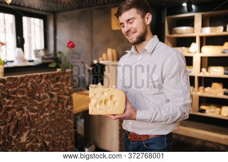 Hansome Man Hold Big Slice Of Cheese Maasdam In Hand. Cheese With Big Holes. Background Of Shelves W