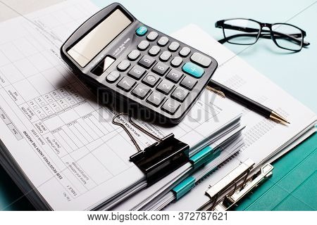 On A Blue Table Are Documents Stacked, A Caculator And Paper Clips