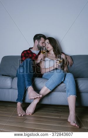 Young Couple Sitting On Sofa Face To Face And Holding Glasses Of White Wine