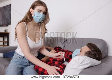Woman In Medical Mask Holding Thermometer Near Sick Boyfriend Lying On Sofa