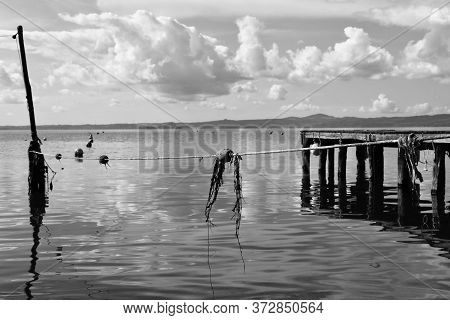 Wooden Jetty For Boats Against A Blue Sky With Some Clouds. Floating Buoys And Fishing Nets. Hills O