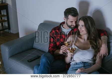Man Embracing Happy Girlfriend While Sitting On Sofa With Glasses Of White Wine