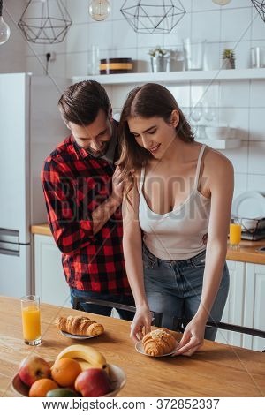 Man Touching Shoulder Of Pretty Girlfriend Holding Plate With Tasty Croissant Near Fresh Fruits