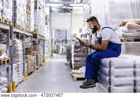 The Storage Worker Is Sitting On The Pile Of Bags With Flour And He Is Using The Tablet For Checking