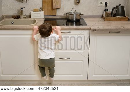 Toddler Baby Climbs On A Hot Electric Stove In The Home Kitchen. A Small Child Touches The Surface O