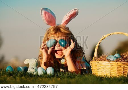 Cute Excited Bunny Child Boy With Rabbit Ears. Child With Easter Eggs In Basket Outdoor. Boy Laying 