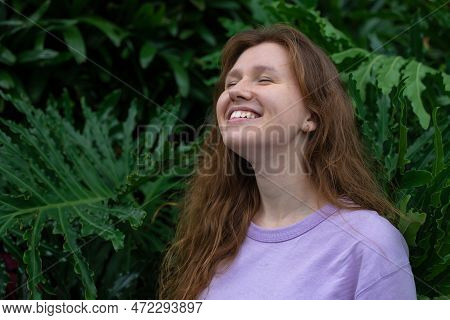 Portrait Of Young Woman Standing On An Ivy, Fern Wall Or In Greenery, Breathing, Smiling And Looking