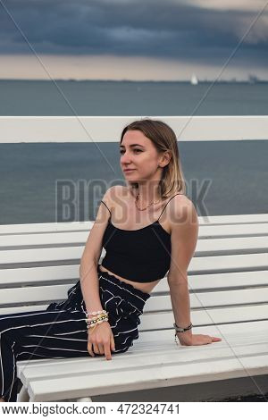 Young Woman Sitting On Bench On Wooden Pier Blurred Beachside Background. Attractive Female Enjoying