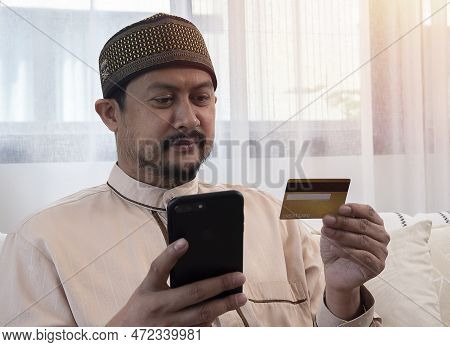 Internet Mobile Shopping Concept. Positive Asian Muslim Man Using Cell Phone Holding Looking At Cred