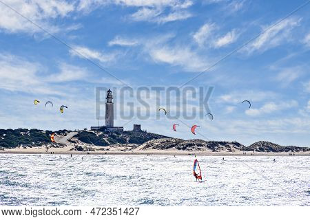 People Practicing Kitesurfing On The Beach Of Los Caños De Meca, Next To The Trafalgar Lighthouse, B