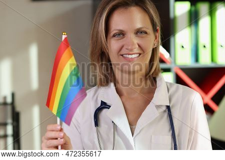 Smiling Female Doctor Holds Rainbow Lgbt Flag In Her Hand In Medical Clinic. Medical Support For Lgb