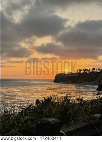 Sun Sets Behind Coastal Bluffs. The Sky Glows In Deep Orange Behind Puff Clouds. High Quality Photo
