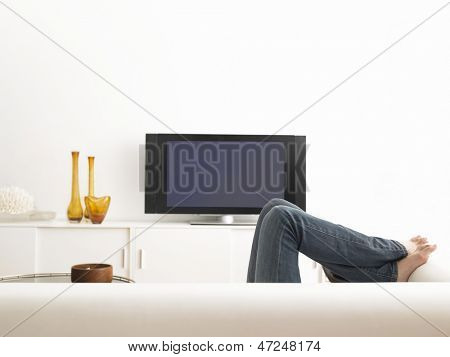 Sección baja de mujer tendida en el sofá con la televisión en la sala de estar contemporánea