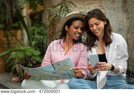 Cheerful Young African American And European Women In Hat Sit On Floor, Looking Route On Map And Sma