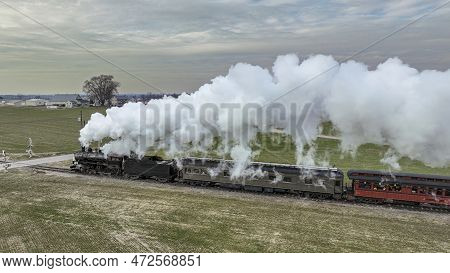A Side Aerial View Of A Steam Passenger Train Approaching, Traveling Thru Open Farmlands, Blowing Lo
