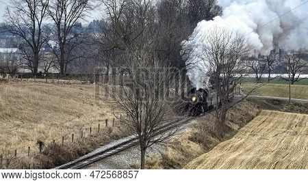 A Drone View Of A Steam Locomotive Approaching Traveling Thru Fields And Meadows, Blowing White Smok