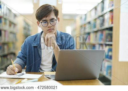 Young Male College Student Wearing Eyeglasses And In Casual Cloths Sitting At Desk Studying, Reading