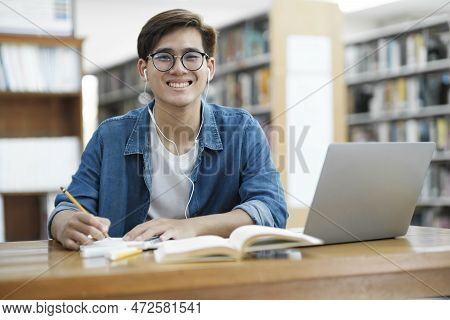 Young Male College Student Wearing Eyeglasses And In Casual Cloths Sitting At Desk Studying, Reading