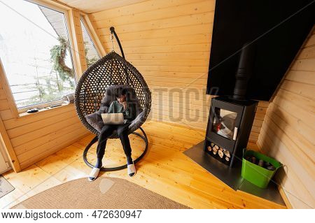 Teenager Boy Sit With Laptop Inside Egg Swing Against Fireplace At Cozy Indoors Heating.