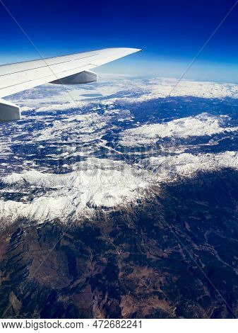 Aerial View Through Porthole Of Plane To Snow-covered Mountain. Scenic View Through Aircraft. Airpla