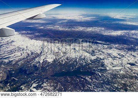 Aerial View Through Porthole Of Plane To Snow-covered Mountain. Scenic View Through Aircraft. Airpla