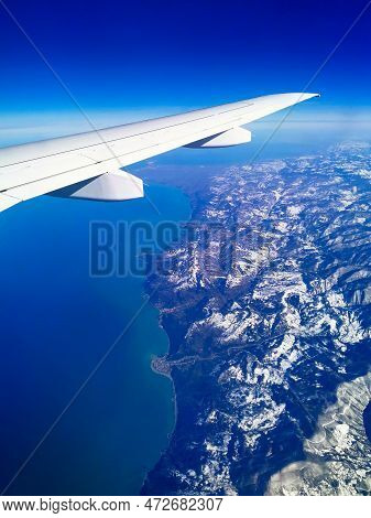Aerial View Through Porthole Of Plane To Snow-covered Mountains And Sea. Scenic View Through Aircraf