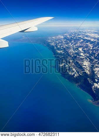 Aerial View Through Porthole Of Plane To Snow-covered Mountains And Sea. Scenic View Through Aircraf