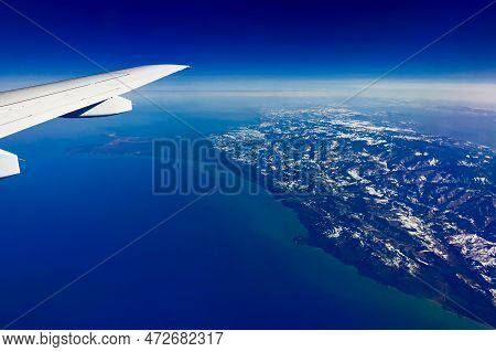 Aerial View Through Porthole Of Plane To Snow-covered Mountains And Sea. Scenic View Through Aircraf