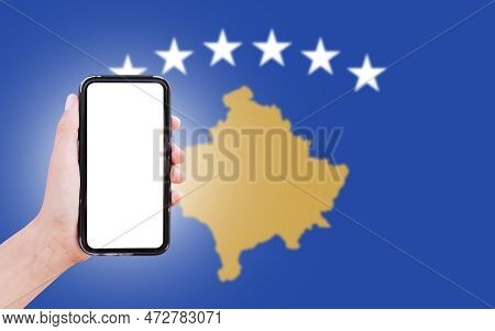 Male Hand Holding Smartphone With Blank On Screen, On Background Of Blurred Flag Of Kosova. Close-up