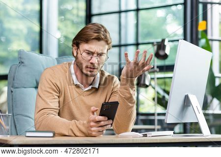 An Angry And Shocked Man Sits In The Office At The Table And Looks Worriedly At The Phone, Spreads H