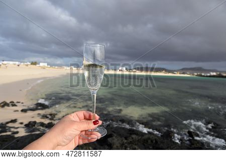 Hand With Glass Of White Champagne Or Cava Sparkling Wine On White Sandy Tropical Beach And Blue Oce