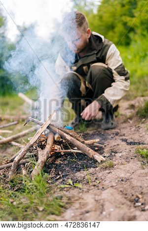 Vertical Shot Of Survivalist Male In Raincoat Starting Fire Using Gas Lighter On Bank Of River For C